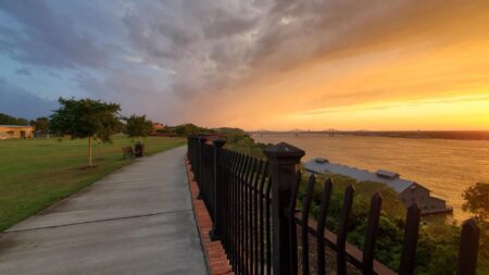 Park with wrought iron fencing next to large body of water with setting sun the background