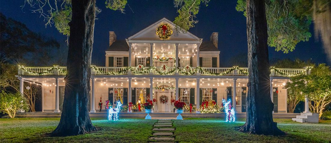 Exterior view of property painted white with green shutters decorated with Christmas decorations and bright lights at night surrounded by green grass and large trees