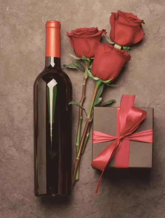 Red wine bottle, three red roses, and small gift wrapped in red ribbon