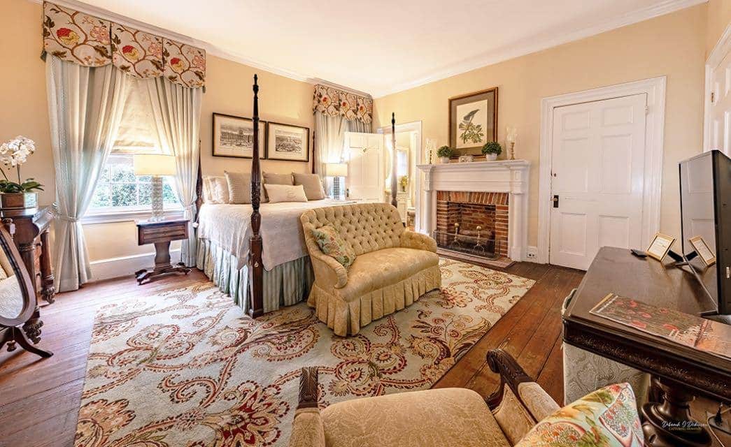 Bedroom with cream walls, white trim, dark hardwood floors, four-poster wooden bed, white and light tan bedding, large windows, and fireplace