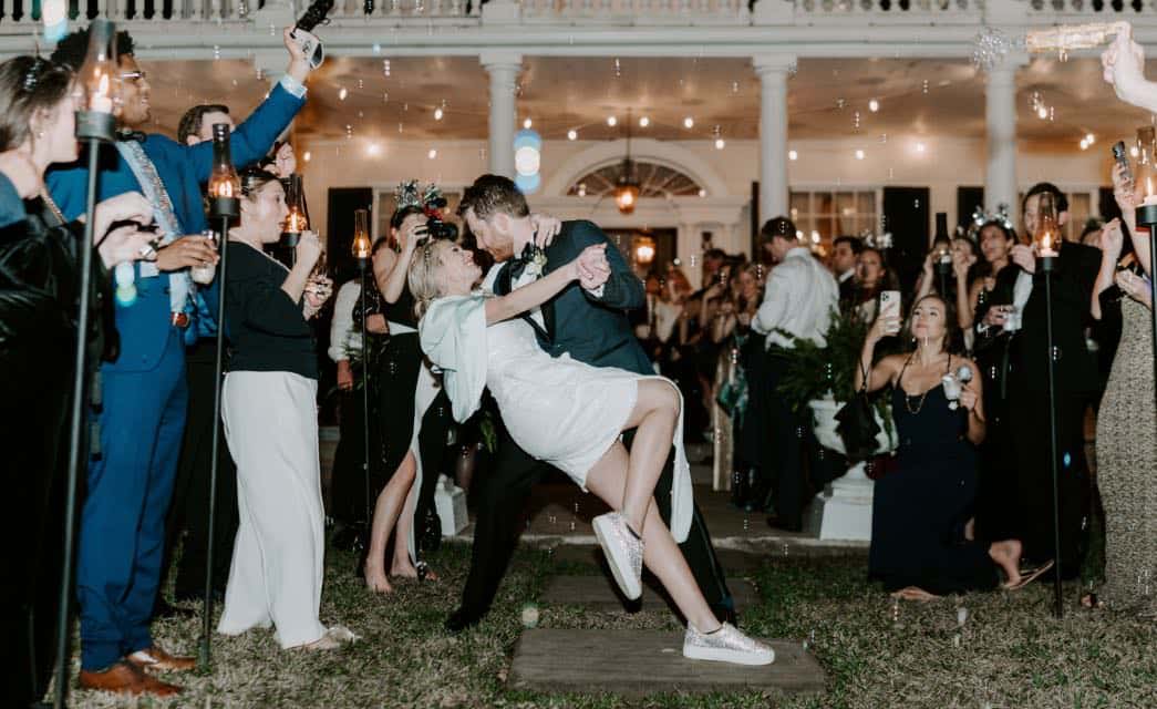 Man in dark suit dipping woman in white dress while people spray bubbles towards the couple