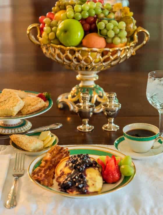 Close up view of plate full of breakfast items next to a cup of coffee, a pastry plate, and large bowl of fresh fruit